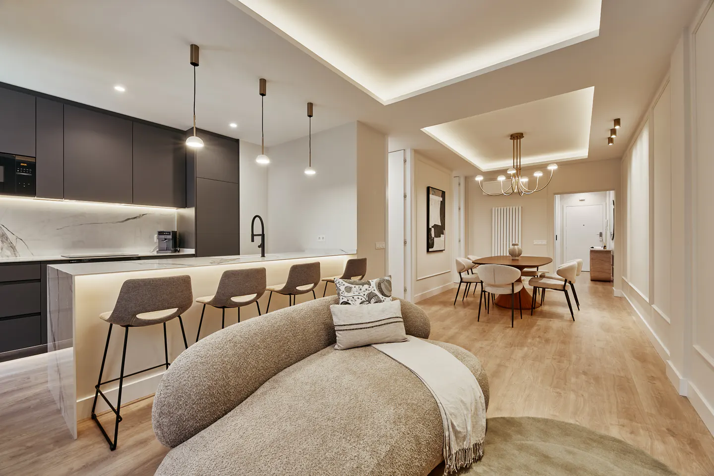 Open-concept living space with a kitchen island, bar stools, and a curved sofa. A dining table and chairs are visible in the background.