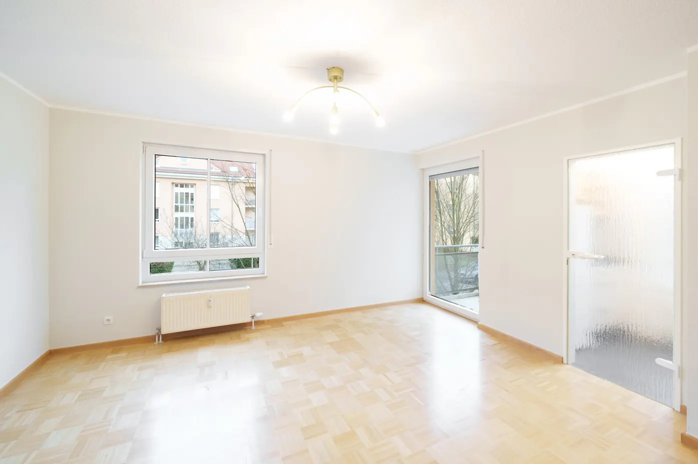 Bright, empty room with parquet floors, white walls, a window, a glass door, and a balcony door.