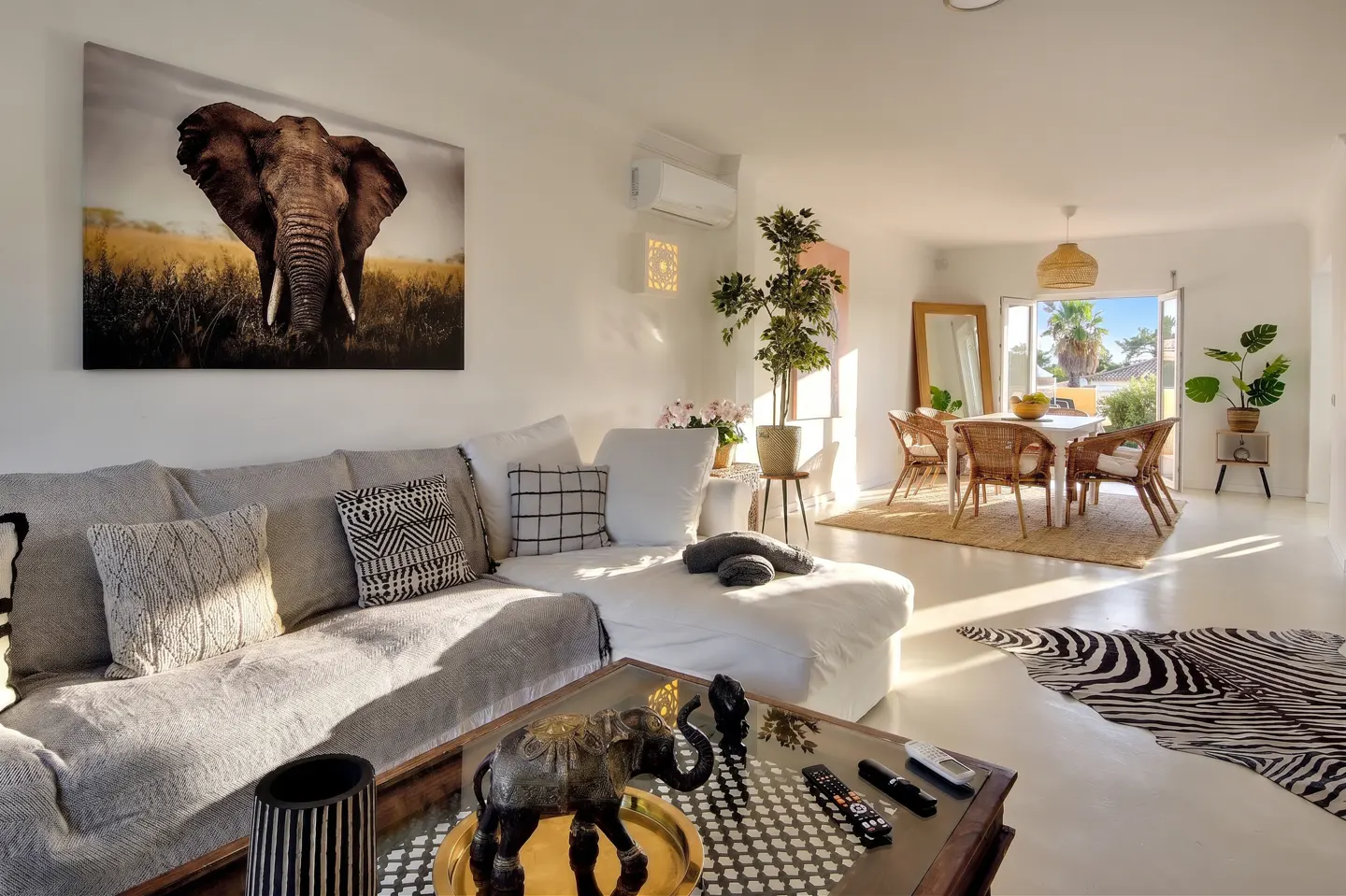 Bright living room with a gray sectional sofa, elephant art, and a dining area with wicker chairs. A zebra-print rug adds a touch of wild style.