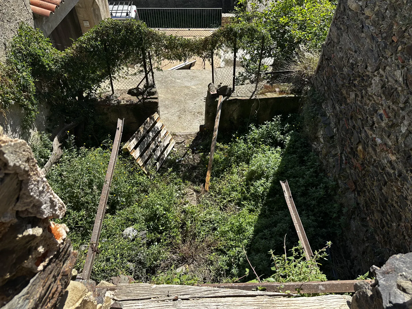 Overhead shot of a neglected yard with overgrown green plants, a wooden pallet, and metal posts, bordered by stone walls.