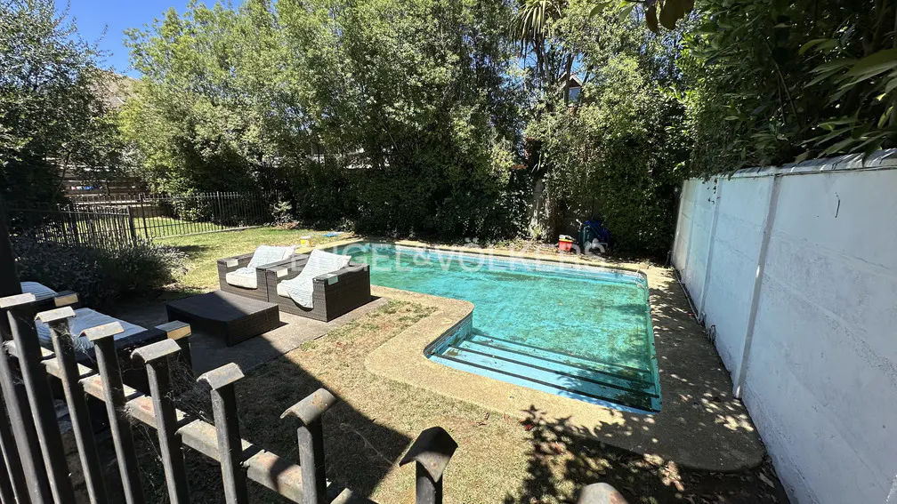 Backyard view of a turquoise swimming pool with patio furniture, surrounded by green trees and a white wall.