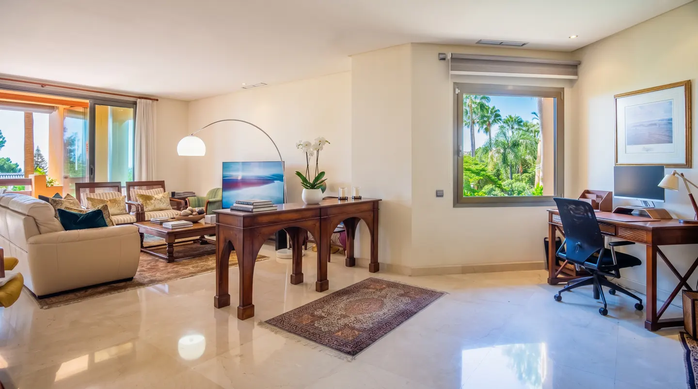 Bright living room with marble floors, a cream sofa, and a wooden desk with a computer by a window overlooking palm trees.