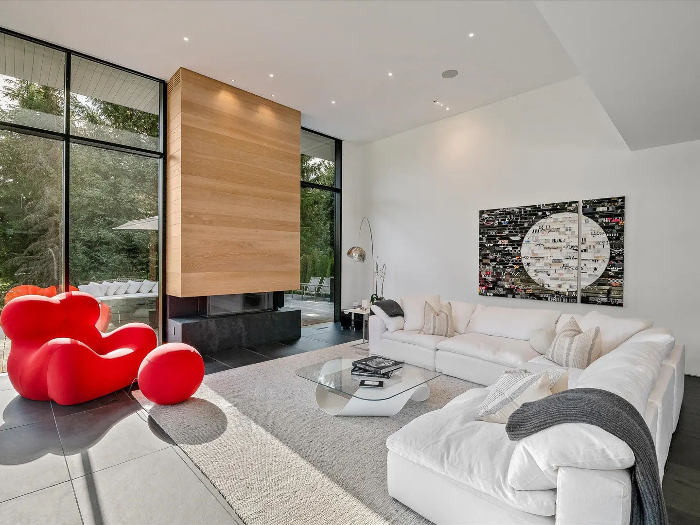 Bright living room with a red chair, white sofa, and a modern fireplace. Large windows offer a view of lush greenery.