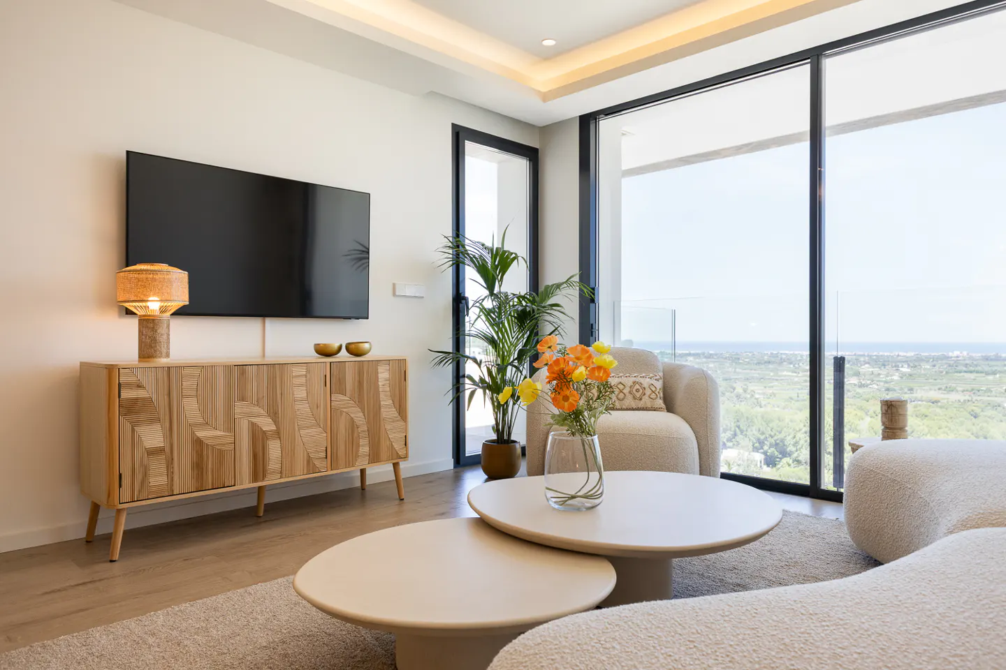 Bright living room with a TV over a wooden cabinet, round tables, and a large window with a view.