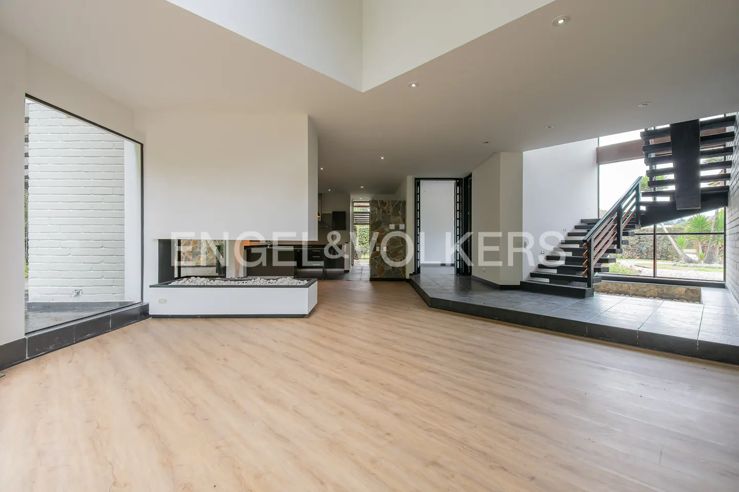 Bright, modern living room with light wood floors, white walls, and a black fireplace. A staircase is visible on the right.