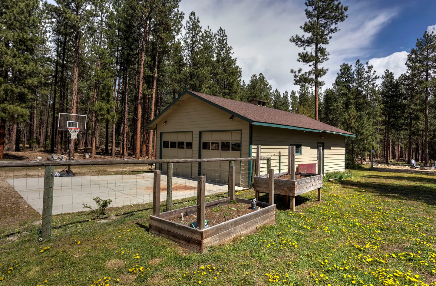 Tan garage with two doors, brown roof, and green trim, surrounded by tall pine trees, a basketball court, and a garden.