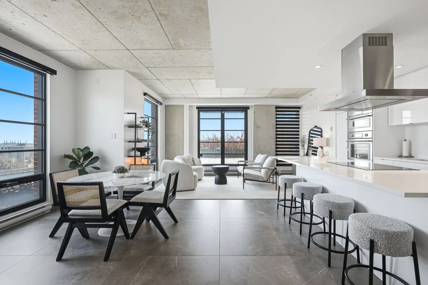 Open-concept condo with a dining table, living room, and kitchen. Large windows offer city views. Gray tile floors and concrete ceilings.
