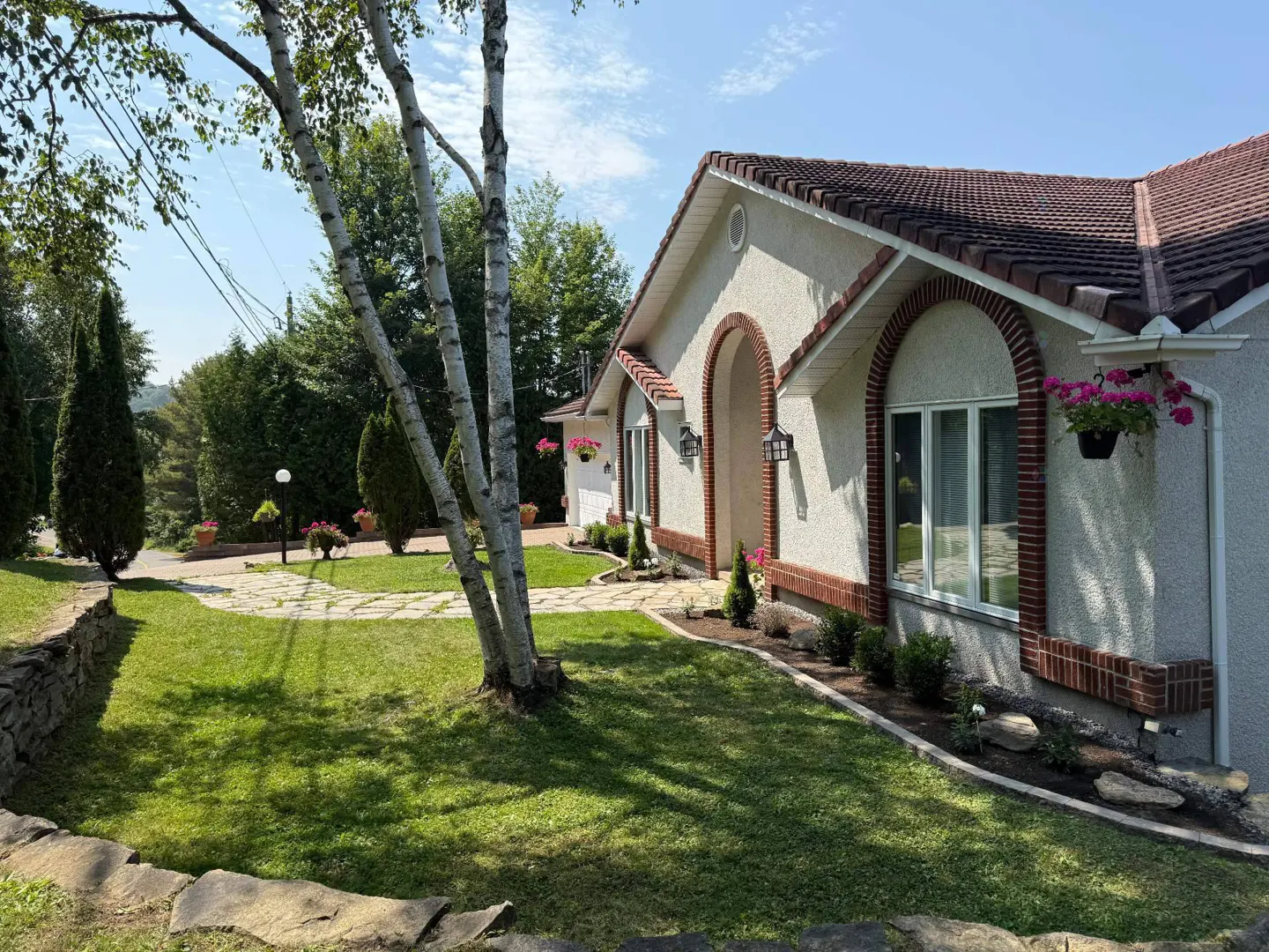 Exterior view of a one-story house with a red tile roof, arched windows, and brick trim. A green lawn and trees surround the house.