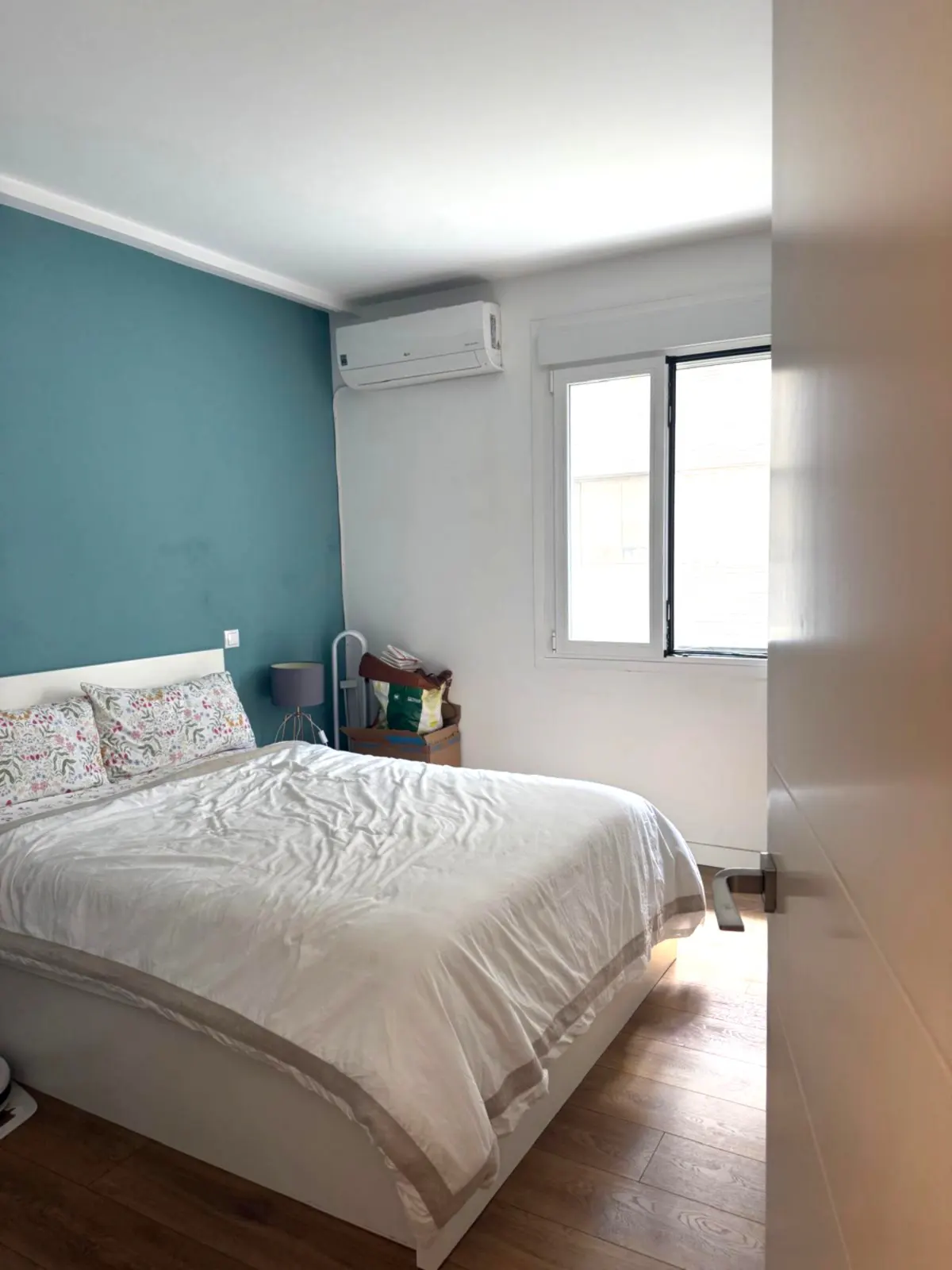 Bright bedroom with a white bed, floral pillows, and a teal accent wall. A window lets in natural light. Wooden floors add warmth.