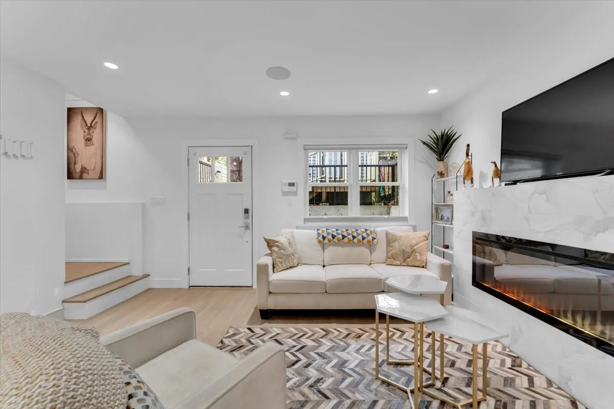 Bright living room with white walls, a beige sofa, a marble fireplace with a TV, and a patterned rug. A white door and stairs are visible.