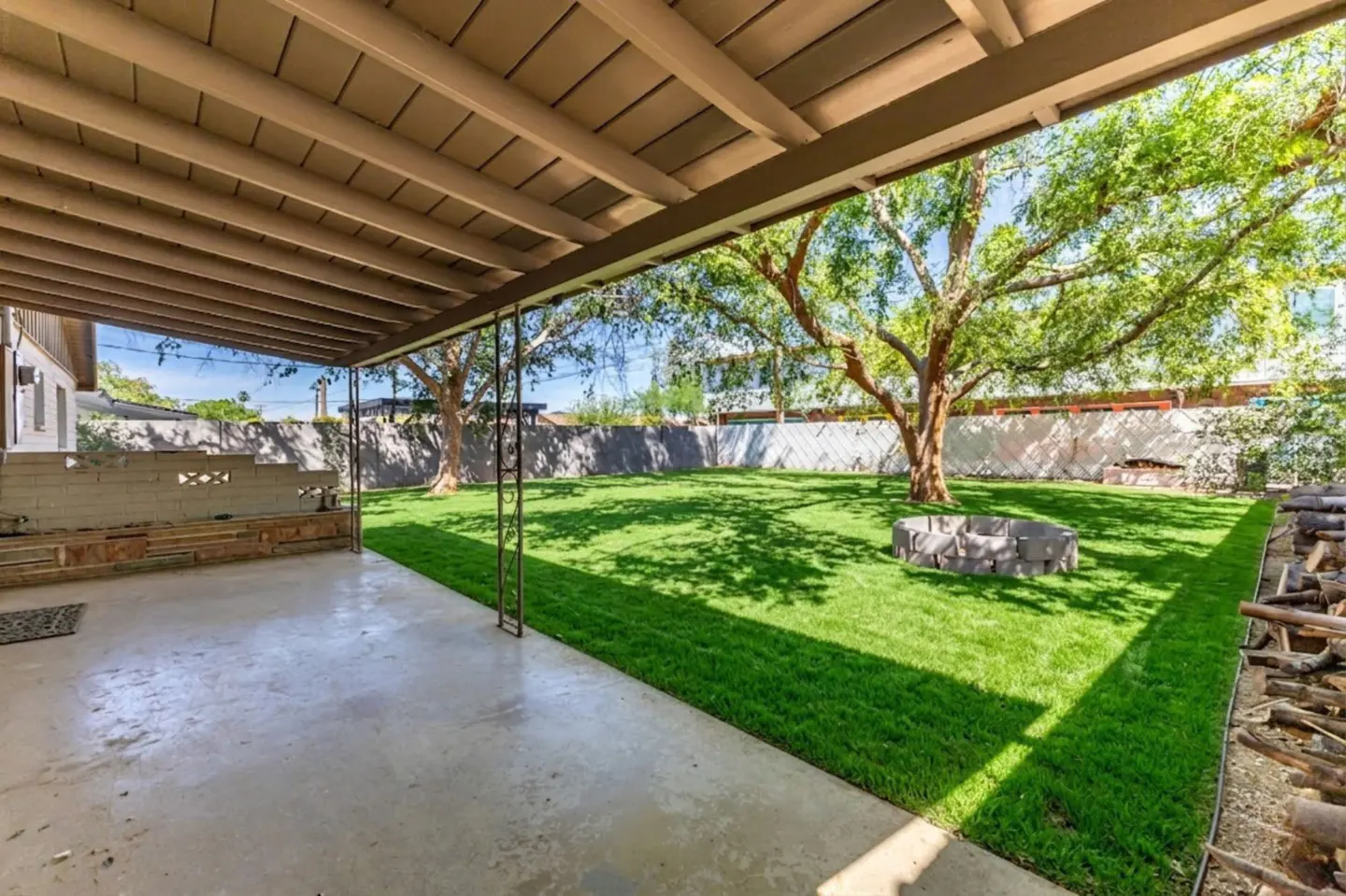Covered patio with a concrete floor leads to a green lawn with trees and a stone fire pit. A white fence surrounds the yard.