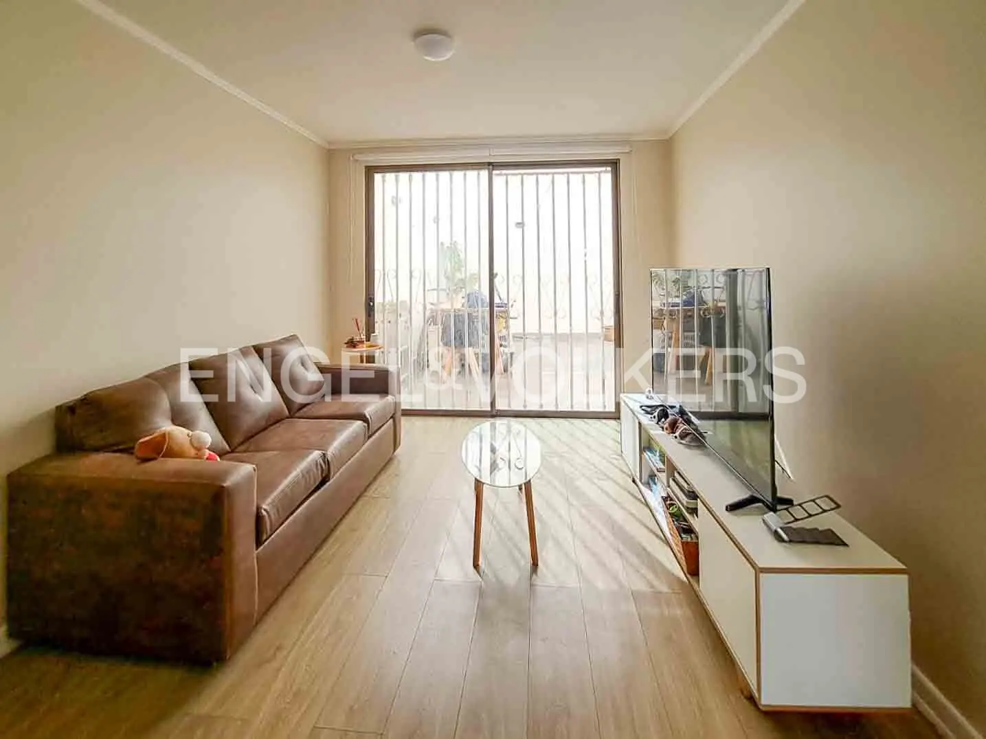 Living room with a brown leather sofa, a glass coffee table, and a TV on a white stand. Sliding glass doors lead to a balcony.