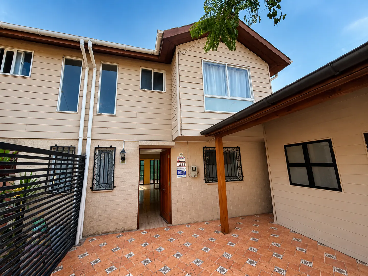 Exterior view of a two-story beige house with a tiled patio, black metal fence, and an open wooden front door.