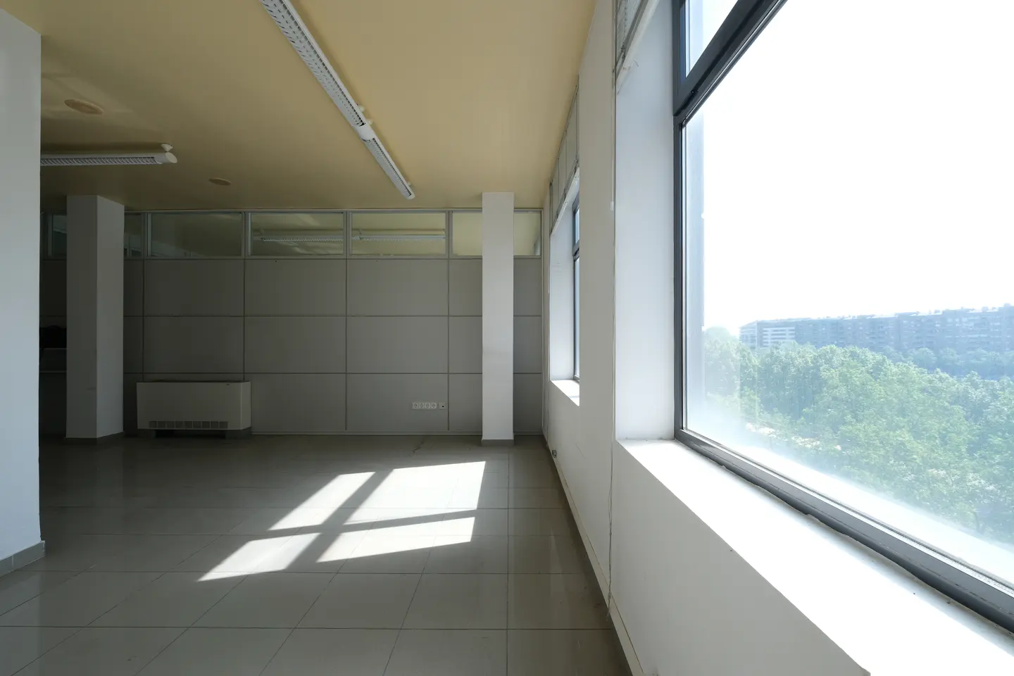 Empty office space with tile floors, white walls, and large windows overlooking a green landscape. Sunlight streams in.