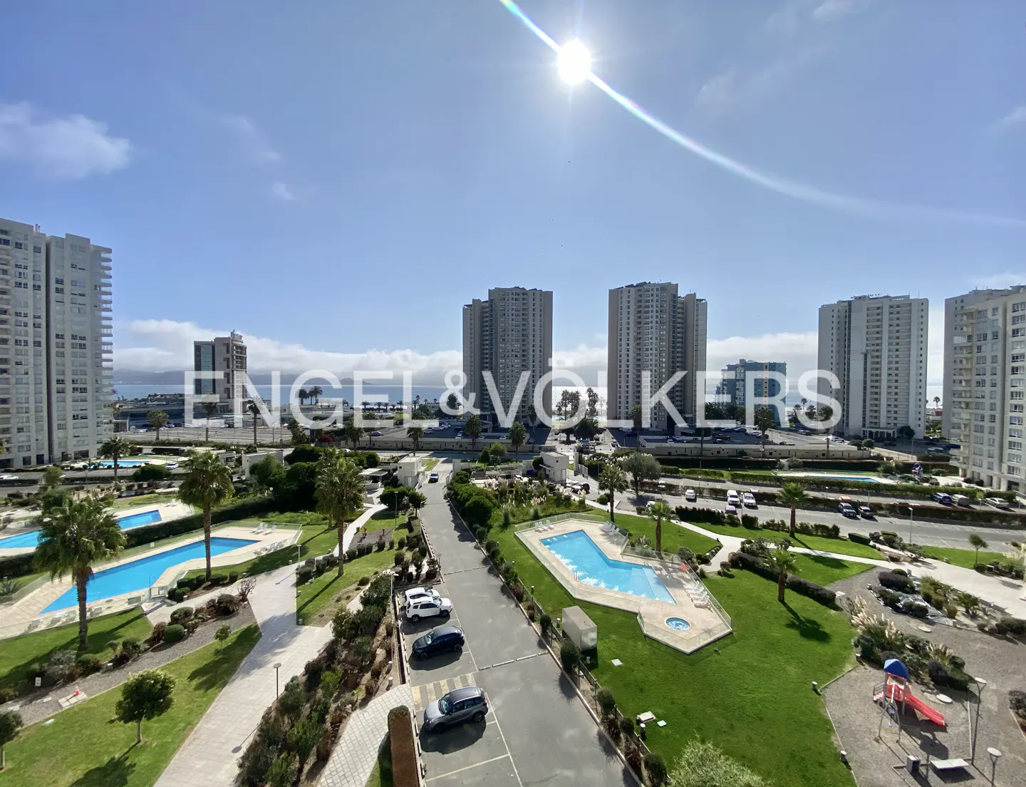 Wide shot of a sunny day overlooking a complex with pools, green spaces, and tall apartment buildings.