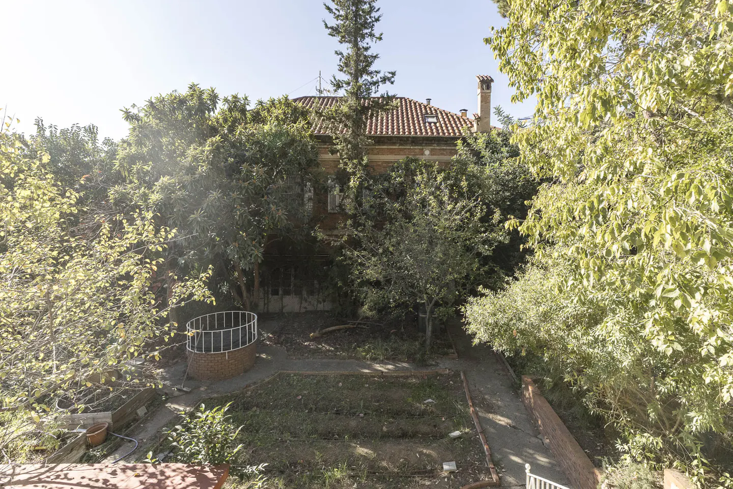 Overhead view of a lush backyard with a brick house, red tile roof, and chimney peeking through trees. A brick well and garden plots are visible.