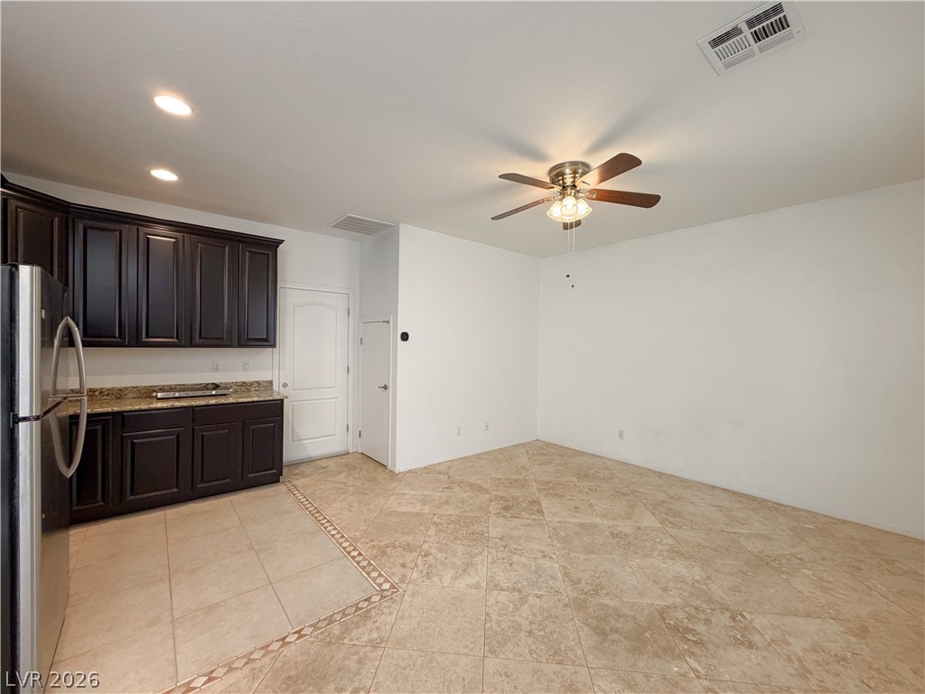 A kitchen with dark wood cabinets, stainless steel refrigerator, and beige tile flooring. A ceiling fan hangs in the center of the room.