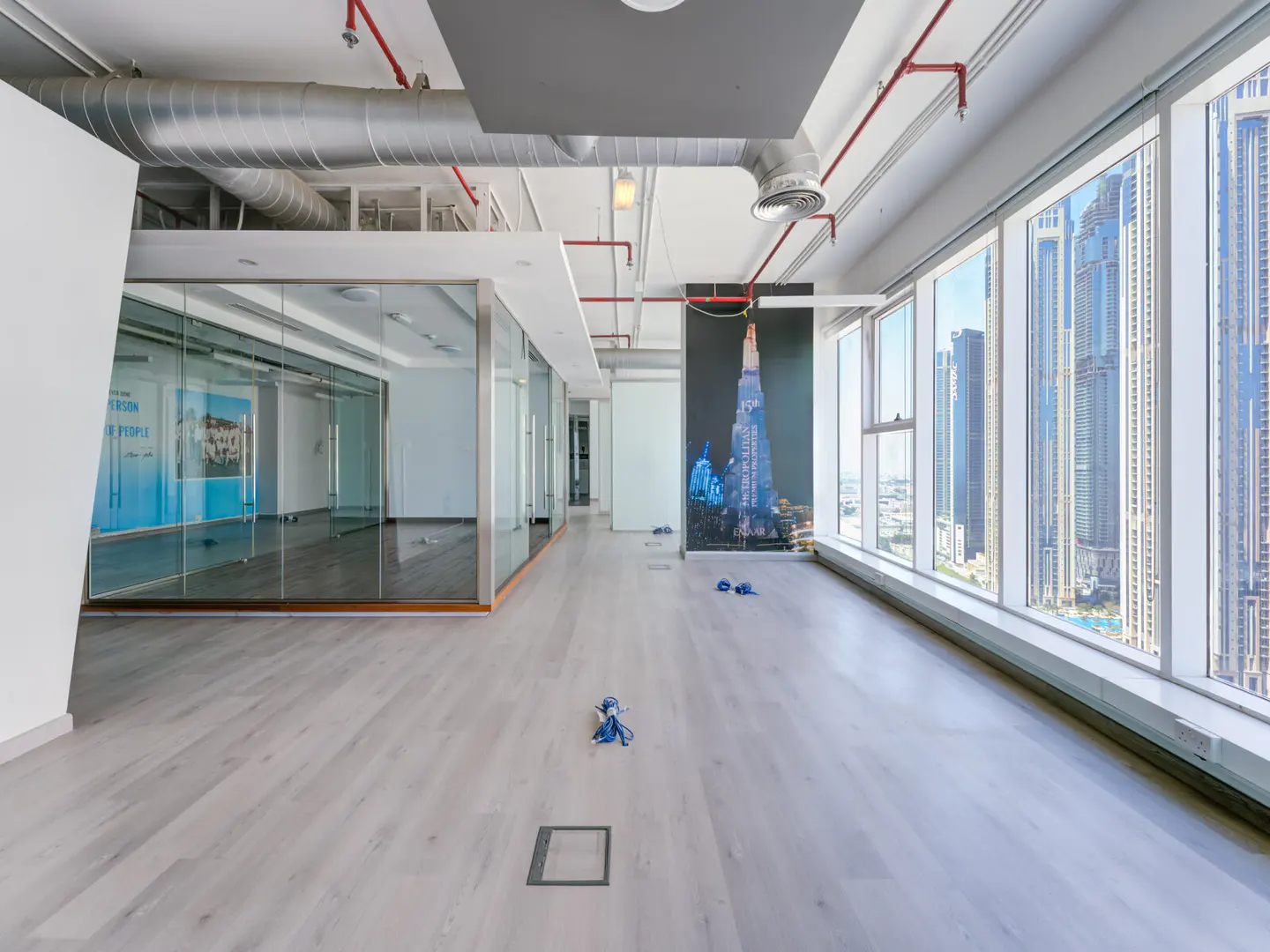 Empty office space with glass-walled rooms, light wood floors, and large windows with city views. Red pipes run along the ceiling.