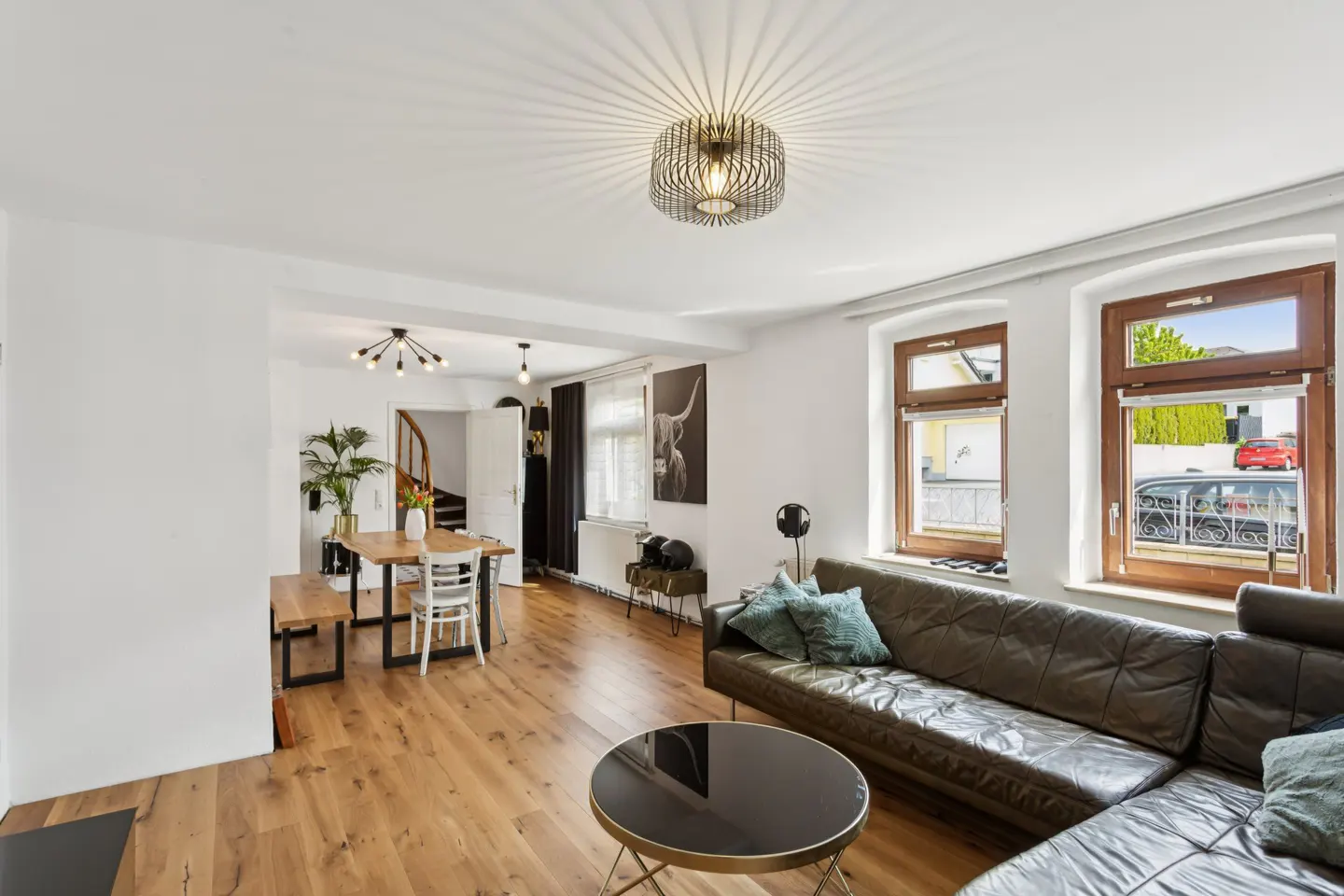 Modern living room with a dark leather sofa, round coffee table, wooden floors, and a dining area with a wooden table.