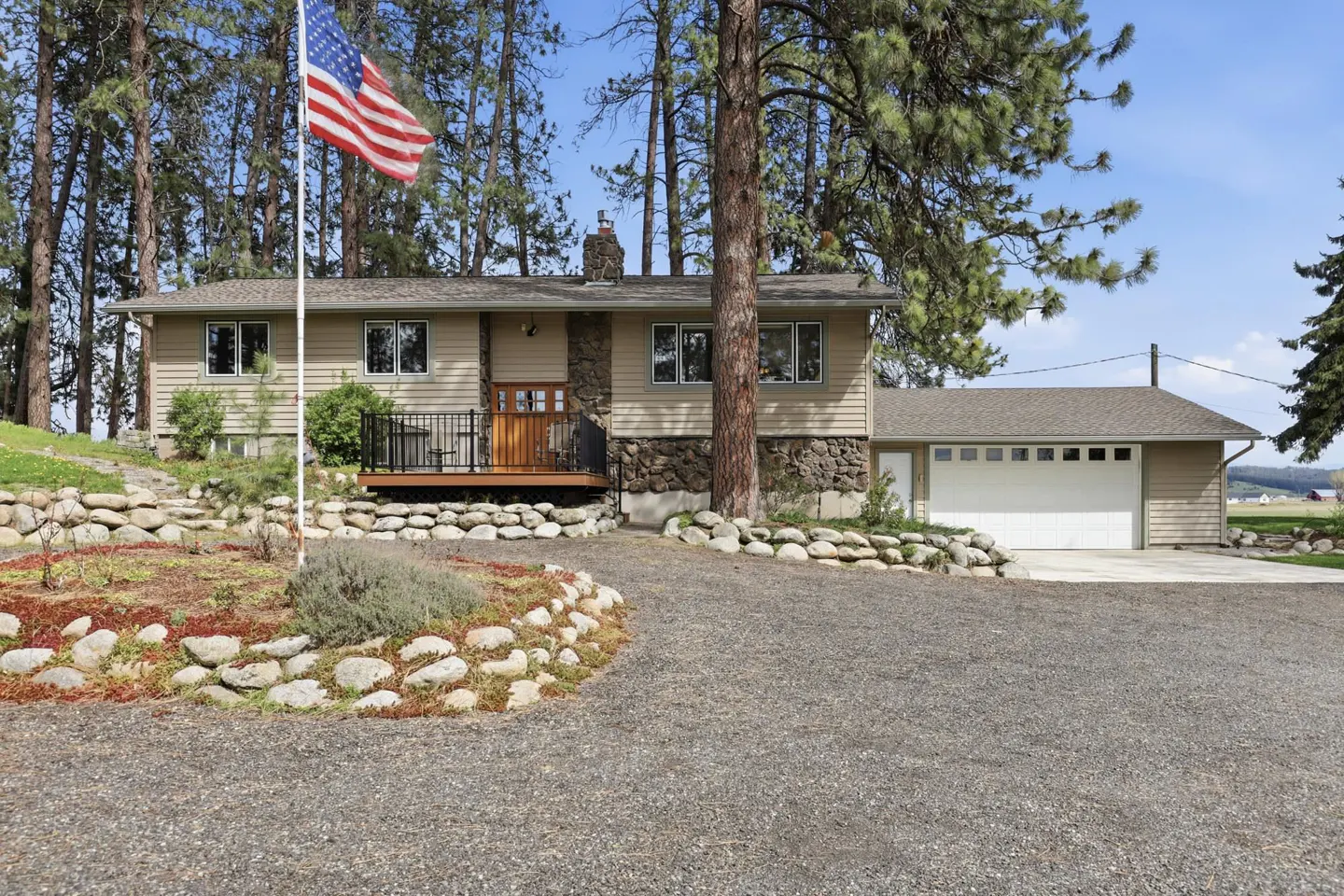 A tan house with a stone facade and an American flag in the front yard. A gravel driveway leads to an attached garage. Tall trees surround the property.