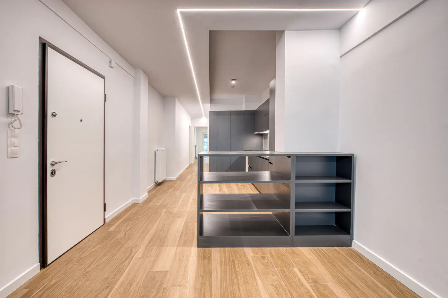 A modern apartment hallway with white walls, wood floors, and a gray shelving unit. A white door with a black frame is on the left.