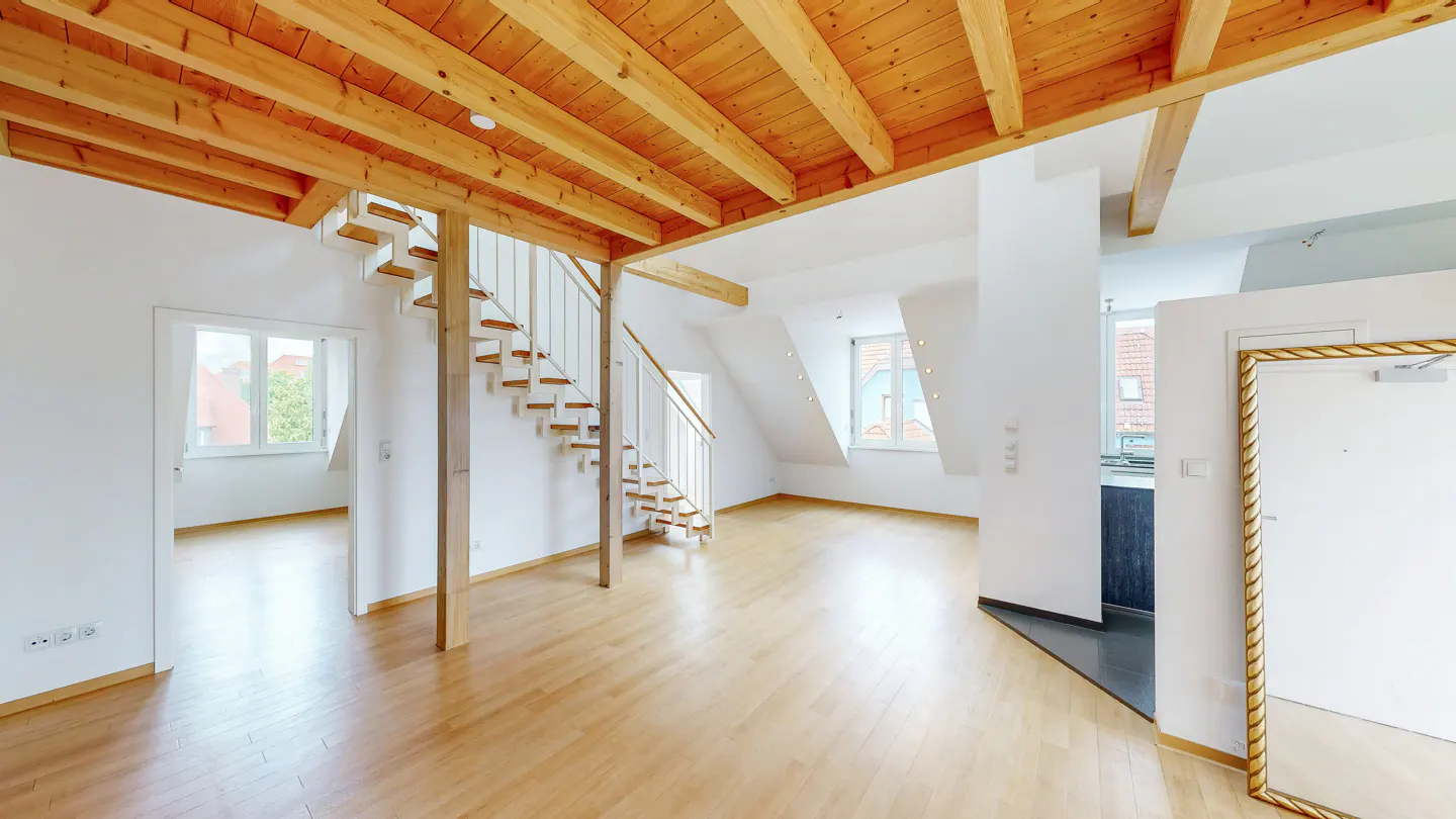 Bright, open-plan attic apartment with light wood floors, exposed wooden ceiling beams, and a modern staircase.
