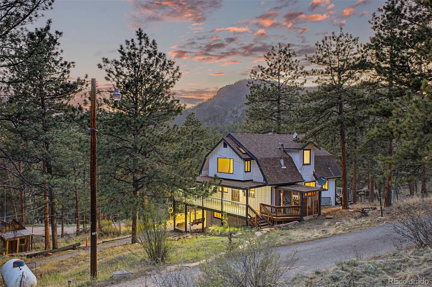 Exterior view of a white house with a brown roof, surrounded by tall pine trees and a mountain backdrop at dusk.