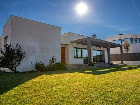 Modern single-story house with a gray pergola, green lawn, and bright blue sky with a shining sun.