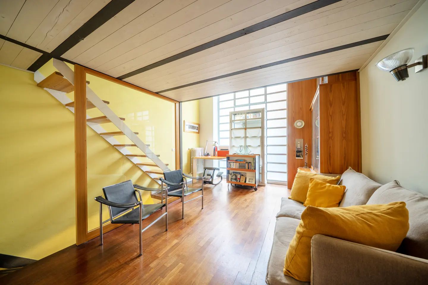 Bright living room with yellow walls, wood floors, and a white staircase. A gray sofa with yellow pillows sits near two black chairs. A desk and bookcase are visible in the background.