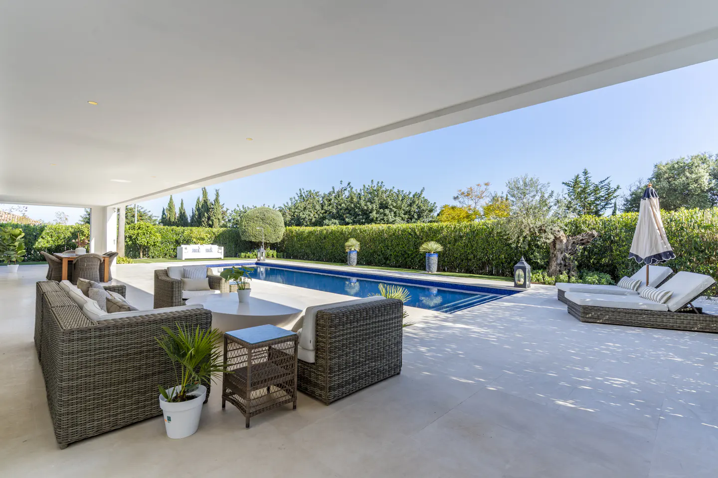 Outdoor patio with wicker furniture, a blue pool, and green hedges under a bright blue sky.