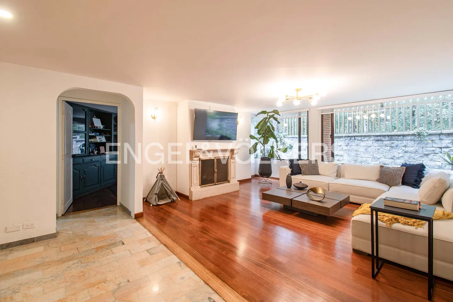 A bright living room with hardwood floors, a white sectional sofa, a fireplace, and a view of the outside.