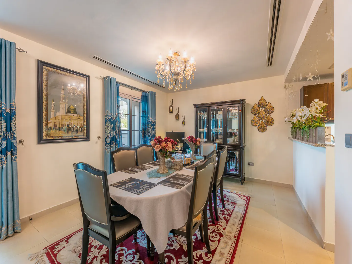 Elegant dining room with a table for six, crystal chandelier, blue curtains, and a framed mosque painting on the wall.
