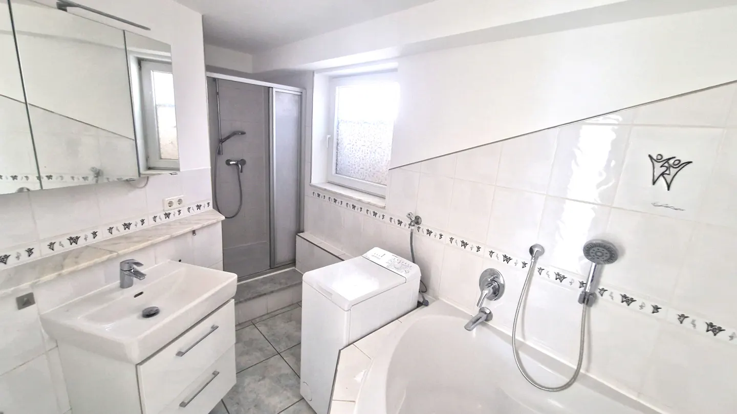 Bright, white bathroom with a sink, shower, tub, and washing machine. The walls are tiled with a decorative border.