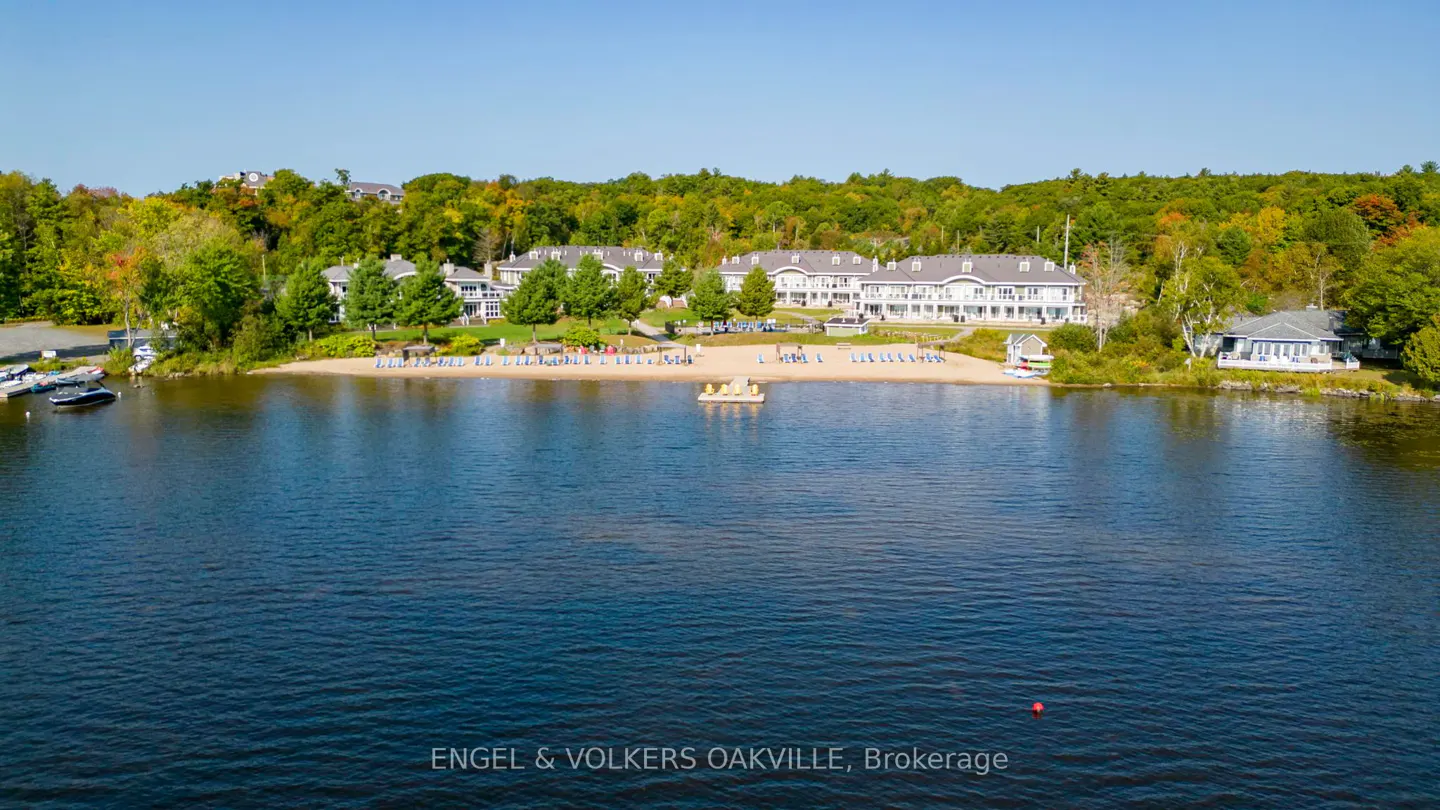 Lakefront property with a sandy beach. White buildings are set back from the beach, surrounded by green trees. Blue water in the foreground.
