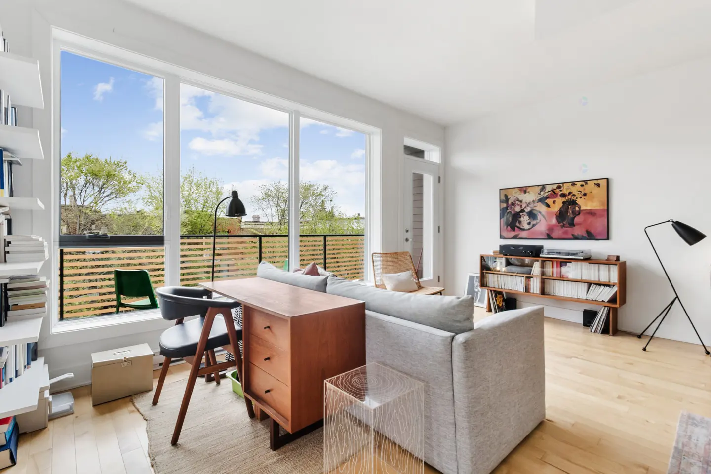 Bright living room with wood floors, large windows, and white walls. A gray sofa faces a wooden media console with a record player. A desk and chair sit by the window.
