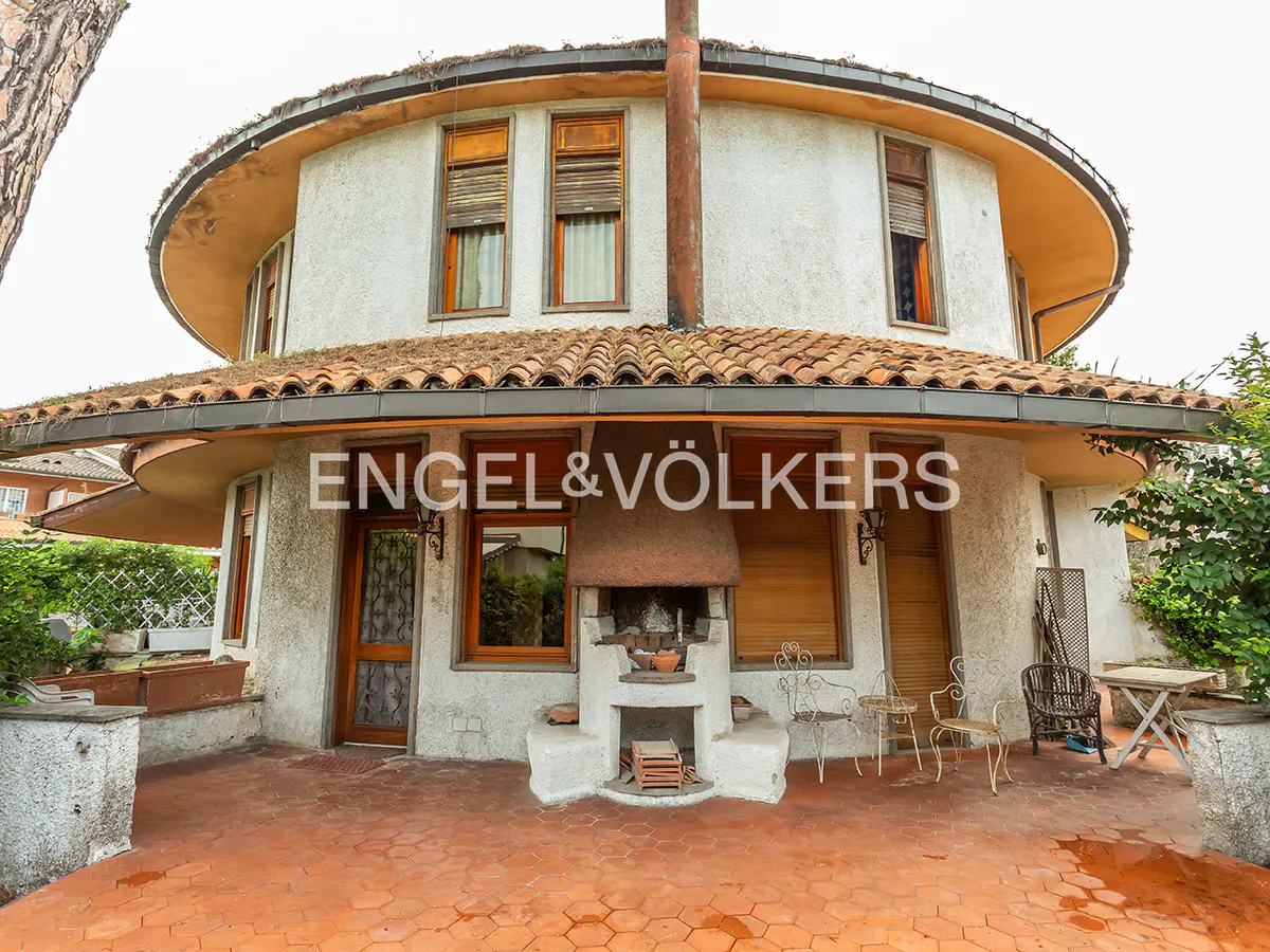 Exterior view of a round, two-story house with a red tile roof and a stone outdoor fireplace. Engel & Völkers logo is visible.