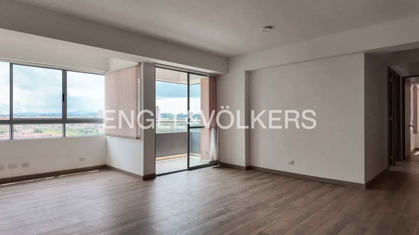Bright, empty room with wood floors, white walls, and large windows overlooking a city. Balcony access. Engel & Völkers logo overlay.