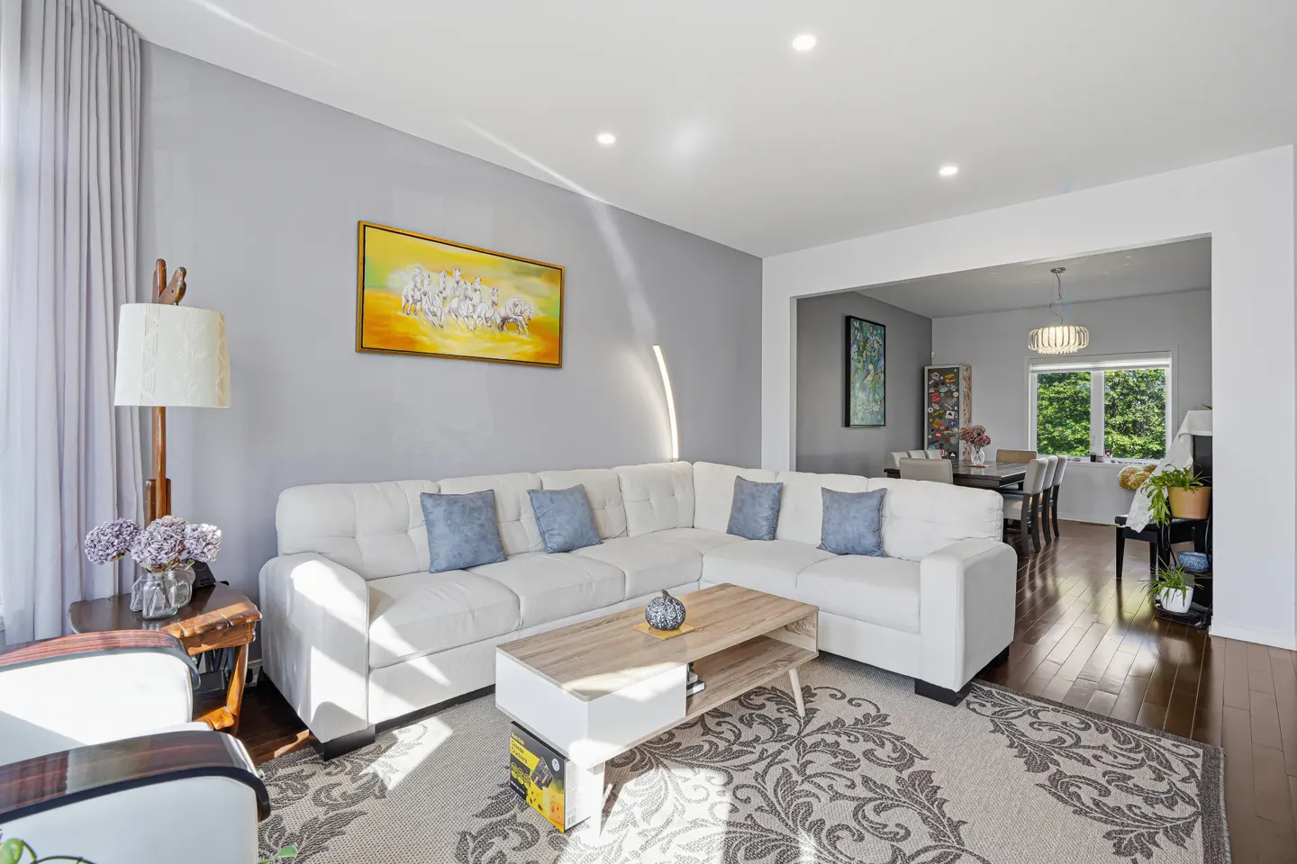 Modern living room with a white sectional sofa, blue pillows, wood coffee table, and a yellow horse painting on gray walls.