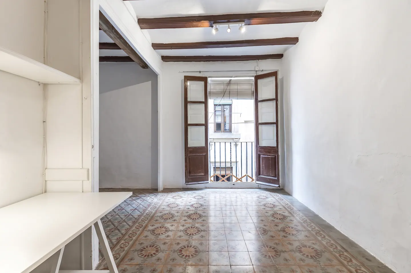Bright room with white walls, patterned tile floor, and open brown doors to a balcony. Wooden beams and lights on the ceiling.