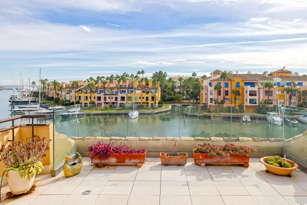 Balcony view of a marina with colorful buildings, boats, and palm trees under a blue sky with white clouds. Planters with plants line the balcony.