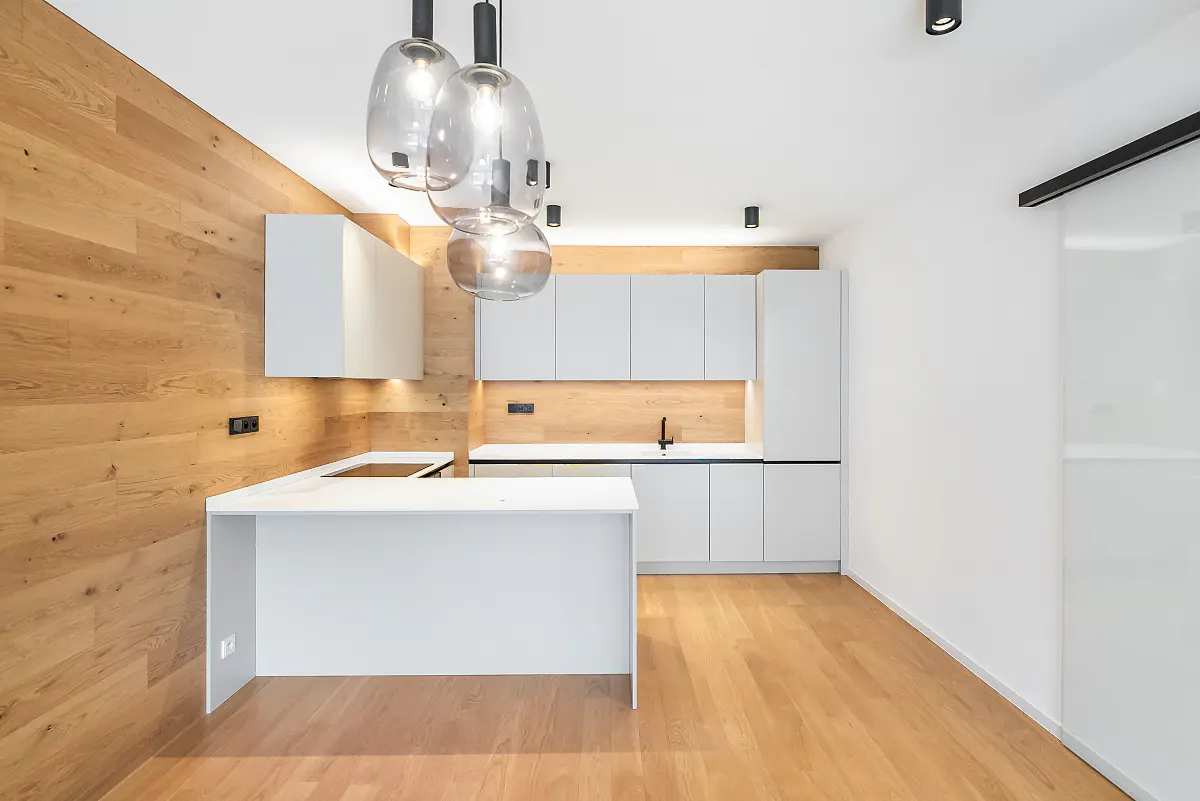 Modern kitchen with white cabinets, a peninsula, and light wood paneling on the walls and floor, lit by glass pendant lights.