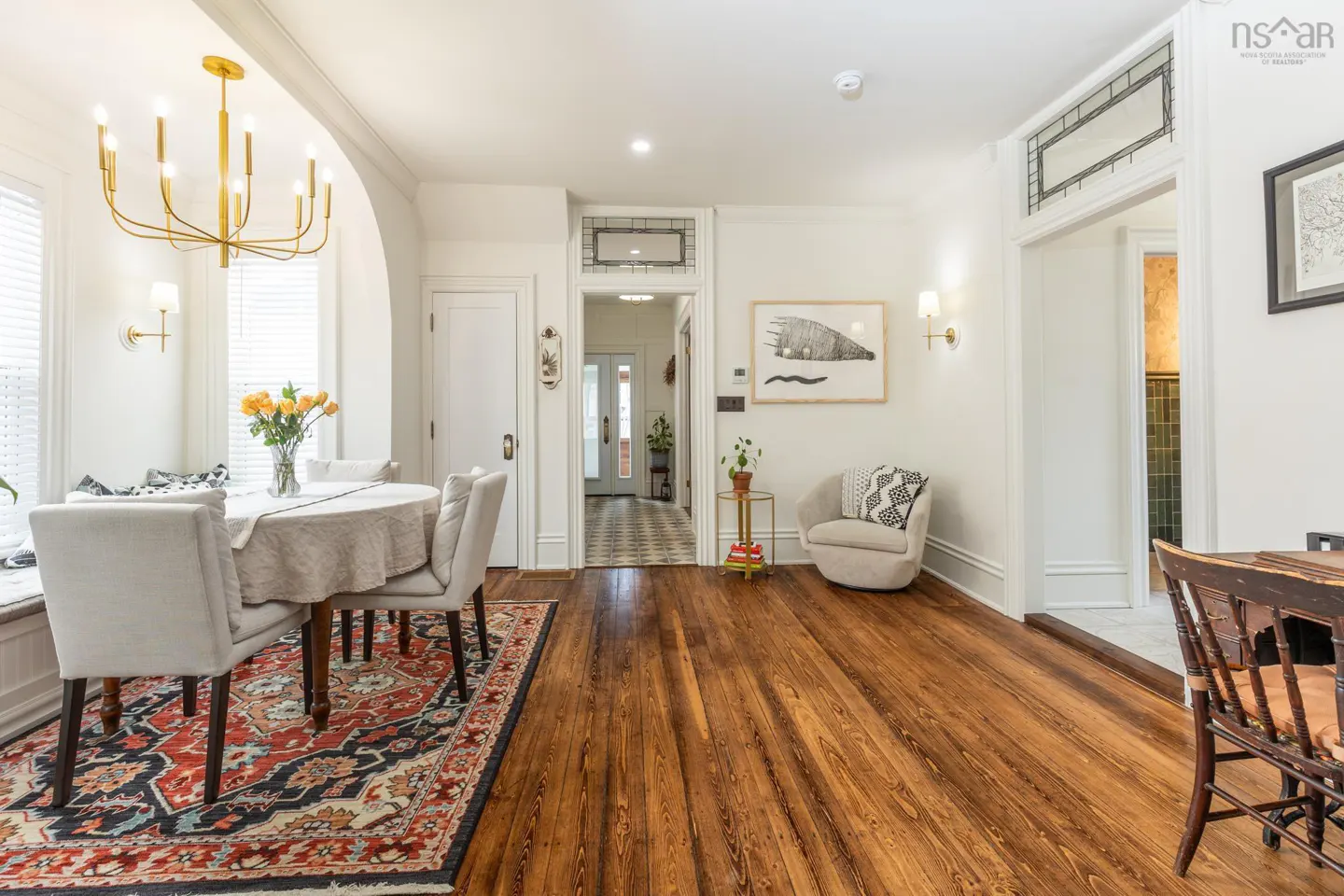 Bright dining room with hardwood floors, white walls, and a gold chandelier. A table with chairs sits on a red patterned rug. Artwork adorns the walls.