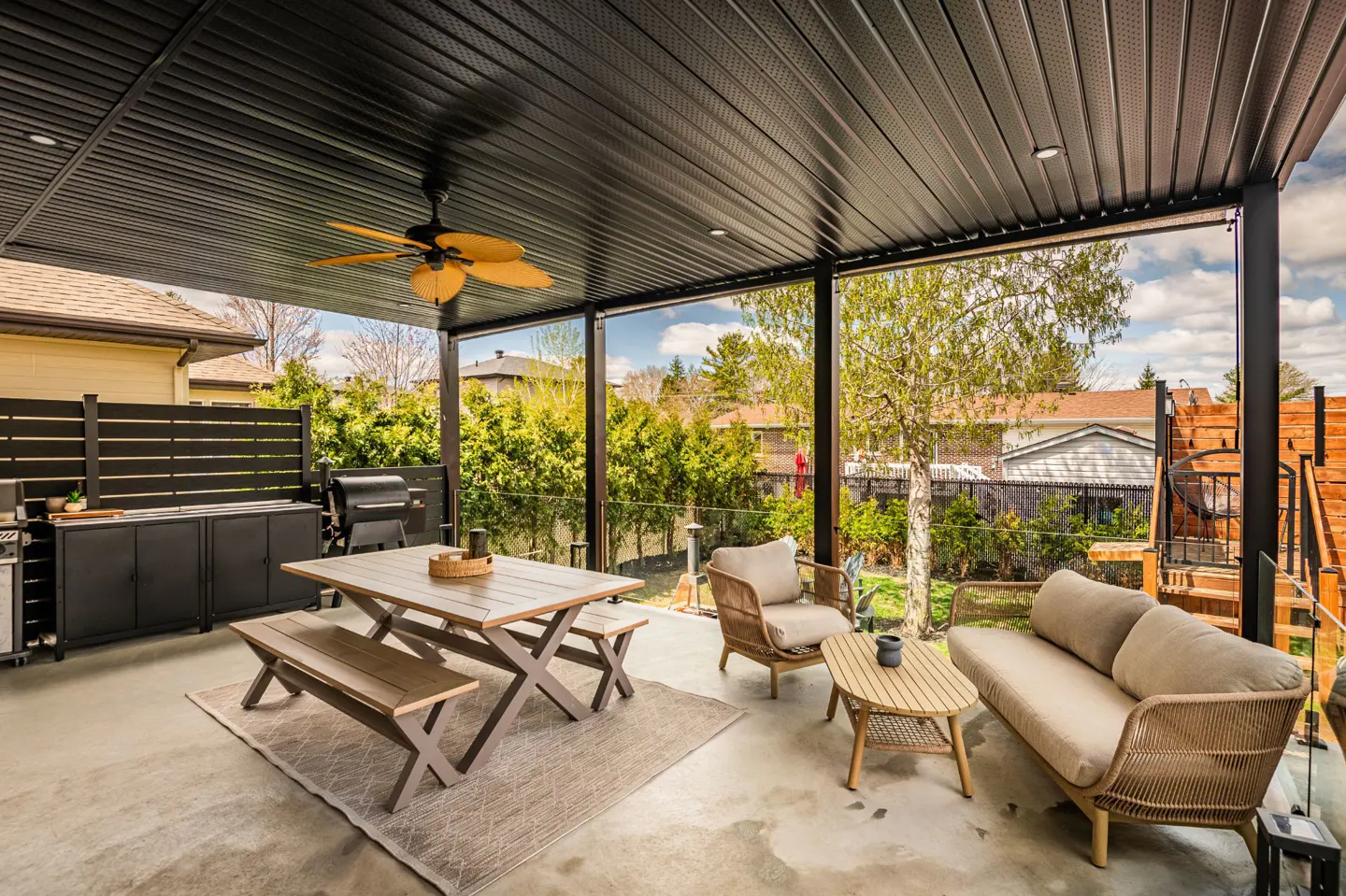 Covered patio with a black ceiling, tan fan, dining table, benches, wicker chairs, and a sofa. A grill and outdoor kitchen are visible.