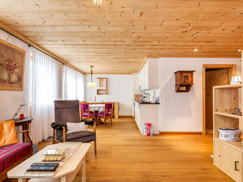 Open-plan living area with wood-paneled ceiling, dining set, cozy armchair, and white kitchen cabinets on hardwood floors.