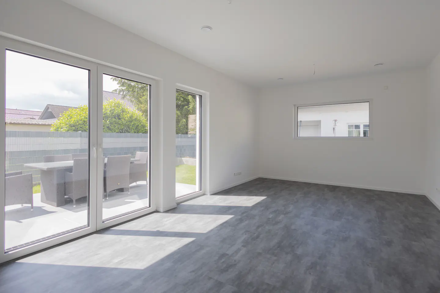 Bright, empty room with gray floors and white walls. Large glass doors lead to a patio with outdoor furniture. A small window is on the right wall.
