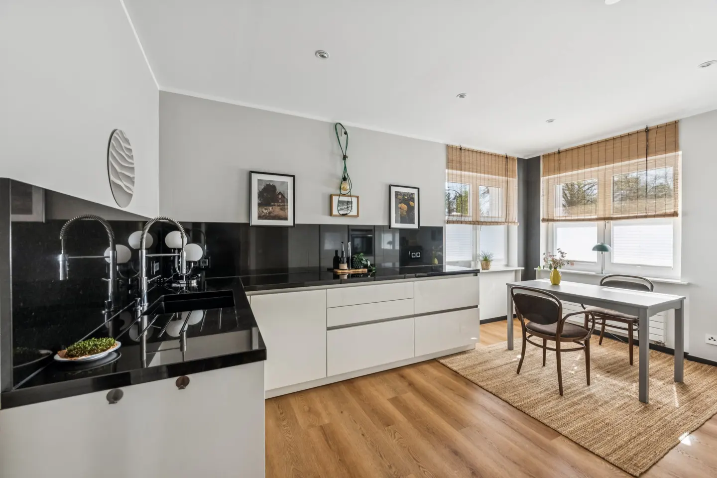 Bright kitchen with black countertops, white cabinets, and a dining table with two chairs on a jute rug. Three windows with bamboo blinds.