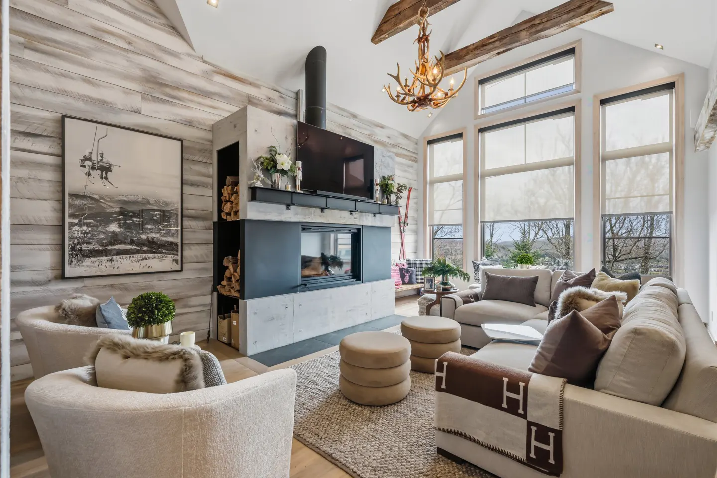 Cozy living room with a fireplace, antler chandelier, vaulted ceilings, and large windows overlooking a wooded landscape.