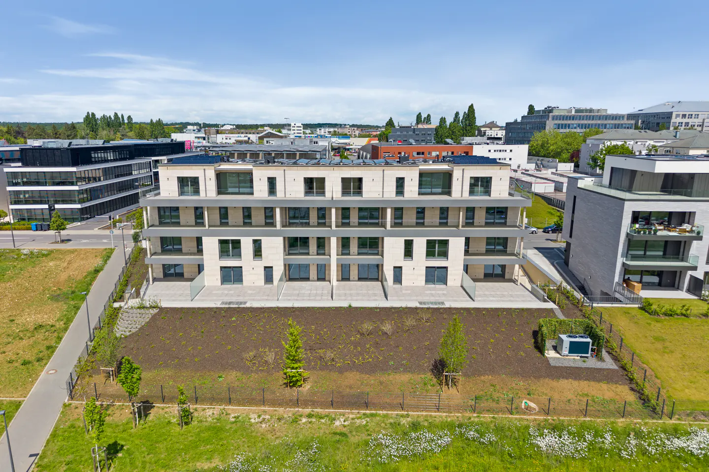 Three-story beige apartment building with large windows and balconies, surrounded by green space and other buildings.