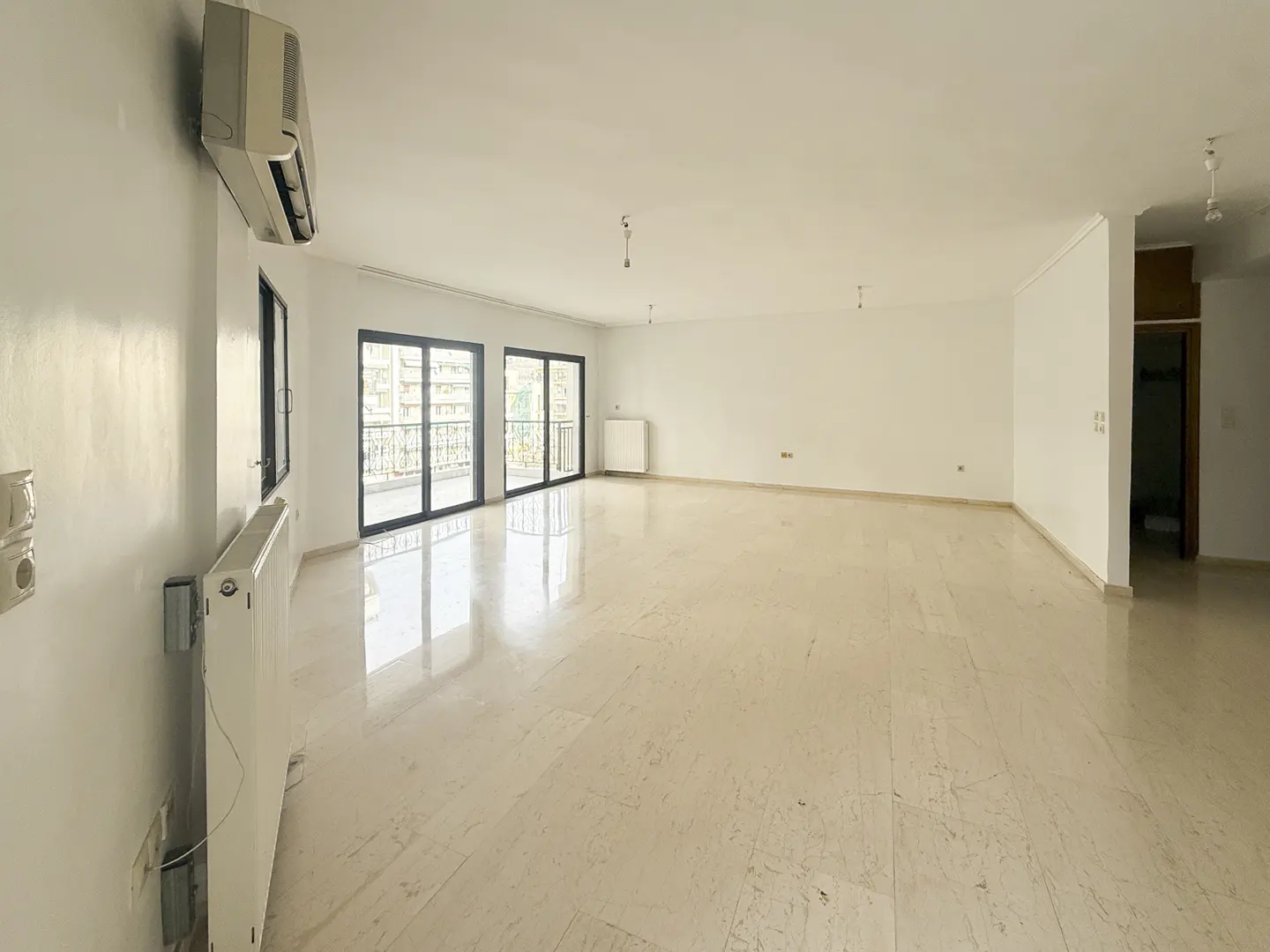 Bright, empty room with marble floors, white walls, and black-framed sliding glass doors to a balcony. Radiators and an air conditioner are visible.