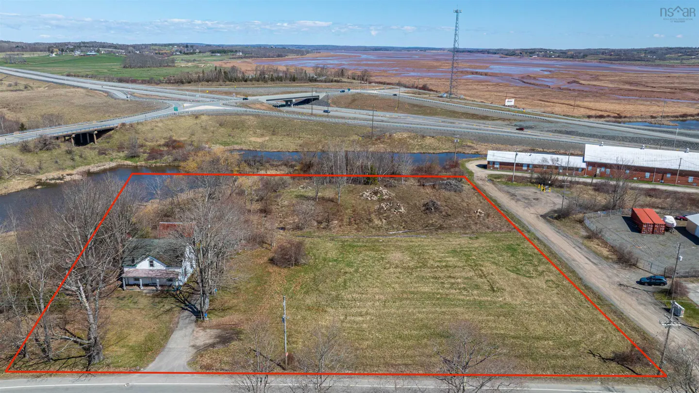 Aerial view of a vacant lot outlined in red, with a small white house, pond, highway, and buildings in the background.